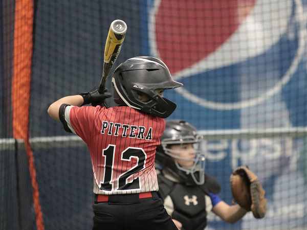Young baseball player in a red jersey with "Pittera 12," ready to bat in an indoor setting. Catcher in gear behind, focused; atmosphere is tense.